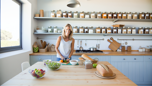 Cocina moderna y luminosa con una mesa de madera llena de alimentos sin gluten, como pan, ensalada de quinoa y frutas. Una mujer caucásica de unos 30 años, vestida de manera casual, sonríe mientras prepara una comida sin gluten. En el fondo, estantes con frascos etiquetados de granos y harinas sin gluten. La escena transmite salud y bienestar, destacando la importancia de la dieta sin gluten en el tratamiento de la enfermedad celíaca.